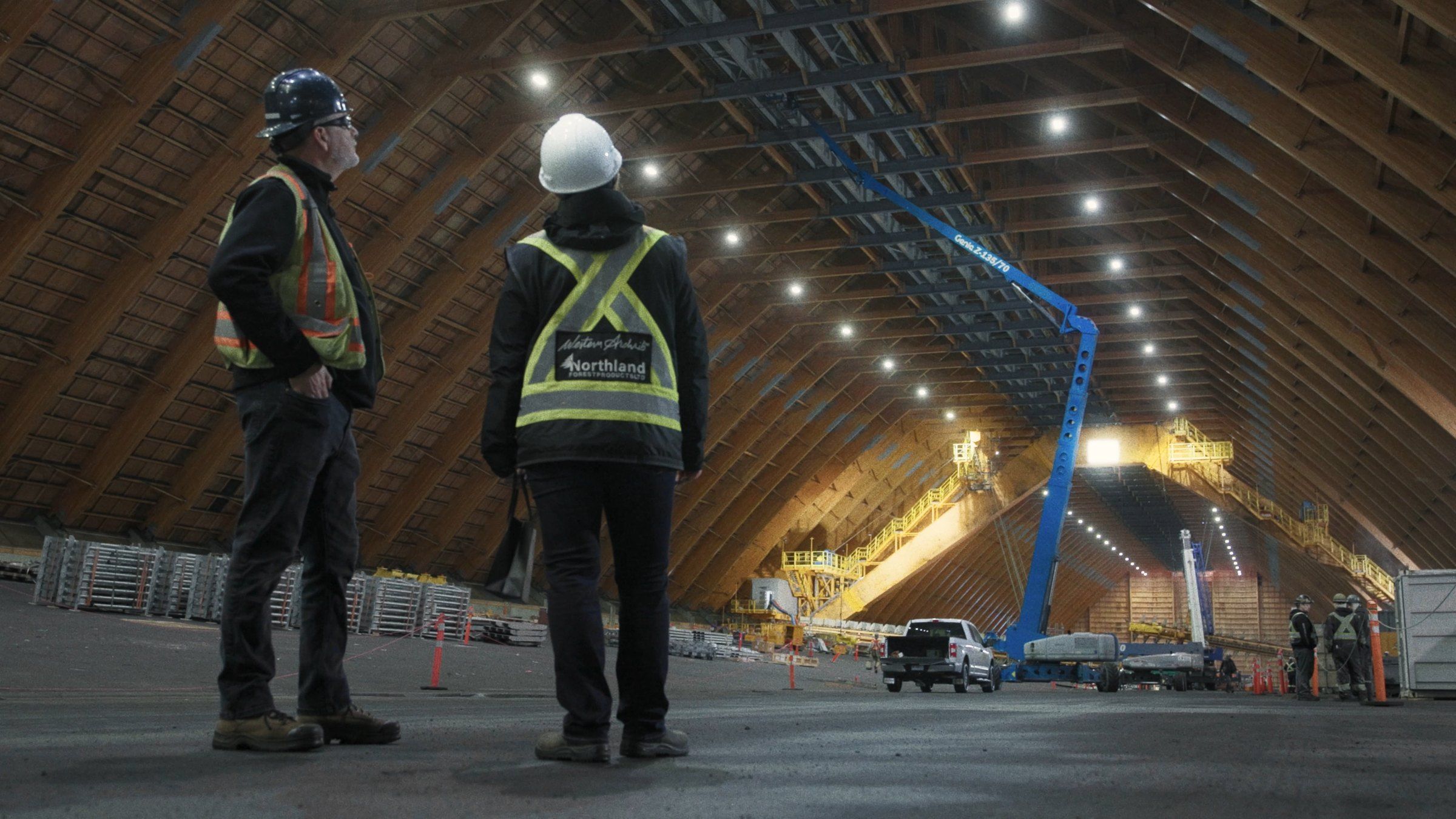 Two people in hard hats look up at the glulam roof from the floor of the building.